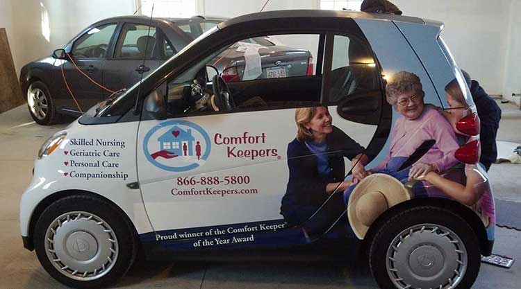 White Smart car wrapped for Comfort Keepers home care, with a caregiver and client photo across the rear panel and a list of services along the door.