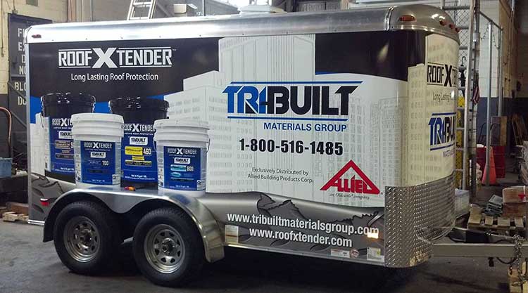 Enclosed cargo trailer wrapped for TRIBUILT Materials Group and RoofXtender, showing product pails against a blue and white skyline graphic.
