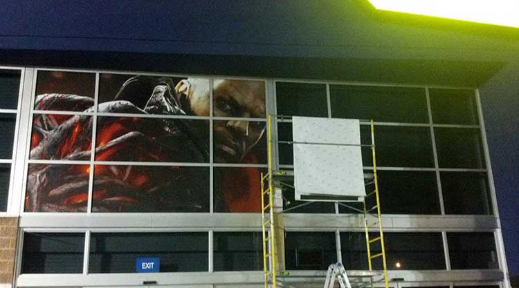 Large exterior window mural being installed across a storefront's second-story glass, with a ladder and scaffolding in place mid-install.