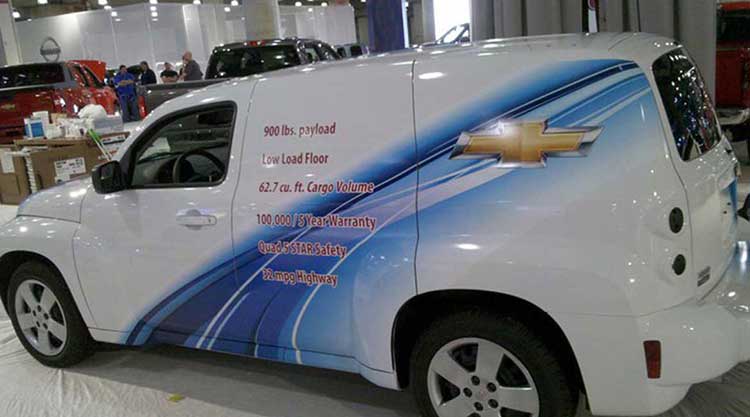 White Chevrolet HHR panel van at an auto show with a blue swoosh graphic and a spec list printed along the side.