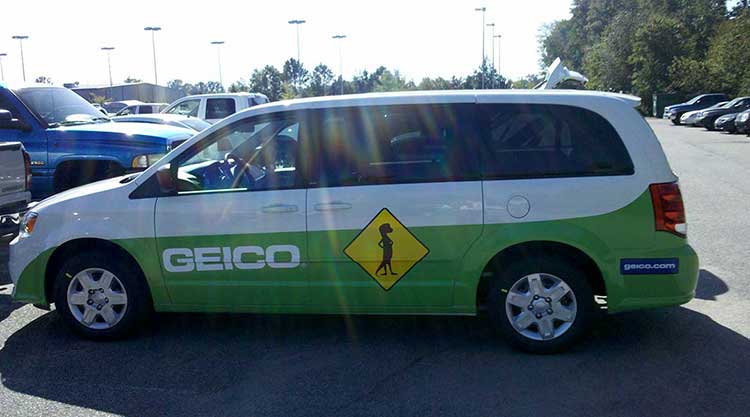 Minivan wrapped in green and white for GEICO, with the logo along the lower body and a crossing-sign graphic on the sliding door.