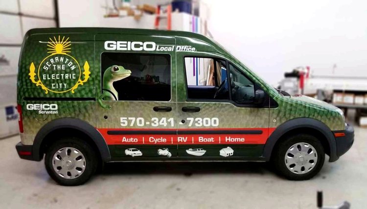 Ford Transit Connect wrapped for the GEICO Scranton local office in green reptile-scale texture, with the gecko peering out of the side window and a Scranton the Electric City crest on the rear panel.